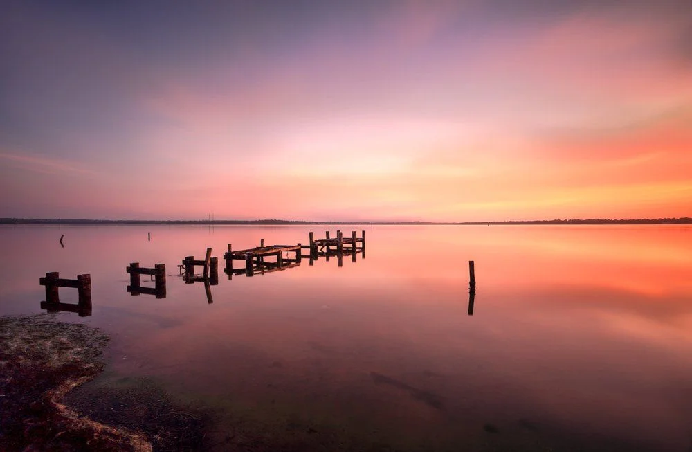 Pink and orange sunset reflected over a calm lake with the remains of an old timber jetty